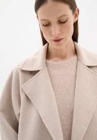 Cappotto in lana beige chiaro con colletto a punta, abbinato a un morbido e leggermente peloso maglione rosa. Entrati i capi presentano un design e una texture minimalista.