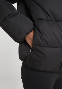 Giacca a vento nera con design trapuntato, dotata di tasca con zip e polsini elastici. Il materiale appare liscio e resistente all'acqua.