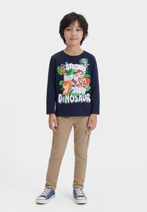Garçon portant une chemise à manches longues bleu marine avec des imprimés colorés de dinosaures et de personnages de dessins animés, un pantalon cargo kaki et des baskets bleues, debout sur fond blanc.