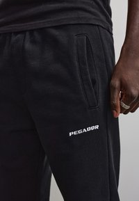 Close-up van een persoon die zwarte joggingbroek draagt met een zijzak en wit PEGADOR-logo, en die zijn rechterhand met een zilveren ring toont.