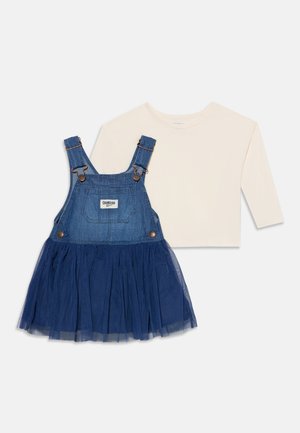Robe salopette en denim avec bretelles boutonnées et jupe en mesh, associée à une chemise crème à manches longues, posée à plat sur fond blanc.