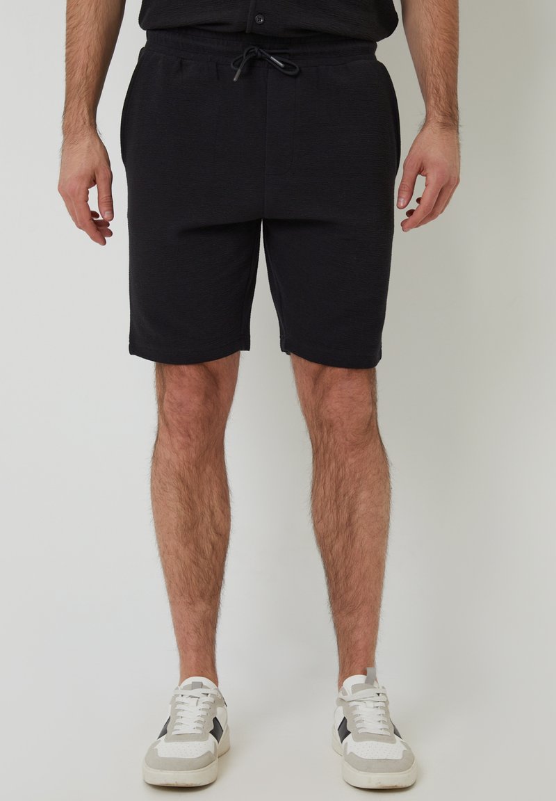Zwarte shorts van een zachte, gestructureerde stof met een elastische tailleband en trekkoord. Gecombineerd met witte sneakers met grijze accenten.