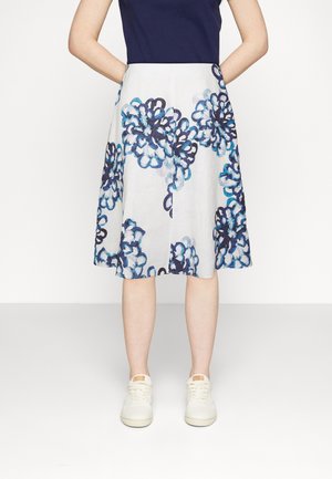 Frau, die einen weißen knielangen Rock mit großen blauen Blumenmustern, ein marineblaues Oberteil und weiße Sneakers trägt, steht mit den Händen hinter dem Rücken.