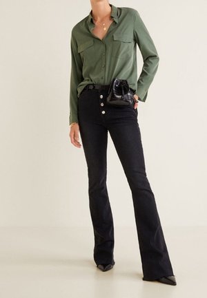 Vrouw draagt een olijfgroen overhemd met knopen, ingestopt in hoge zwarte wijde jeans met een kleine zwarte crossbodytas.
