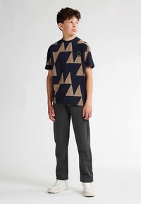Marineblaue T-Shirt mit kurzen Ärmeln, beigen geometrischen Mustern und einem kleinen runden Akzent, kombiniert mit dunkelgrauer Hose und weißen Turnschuhen.