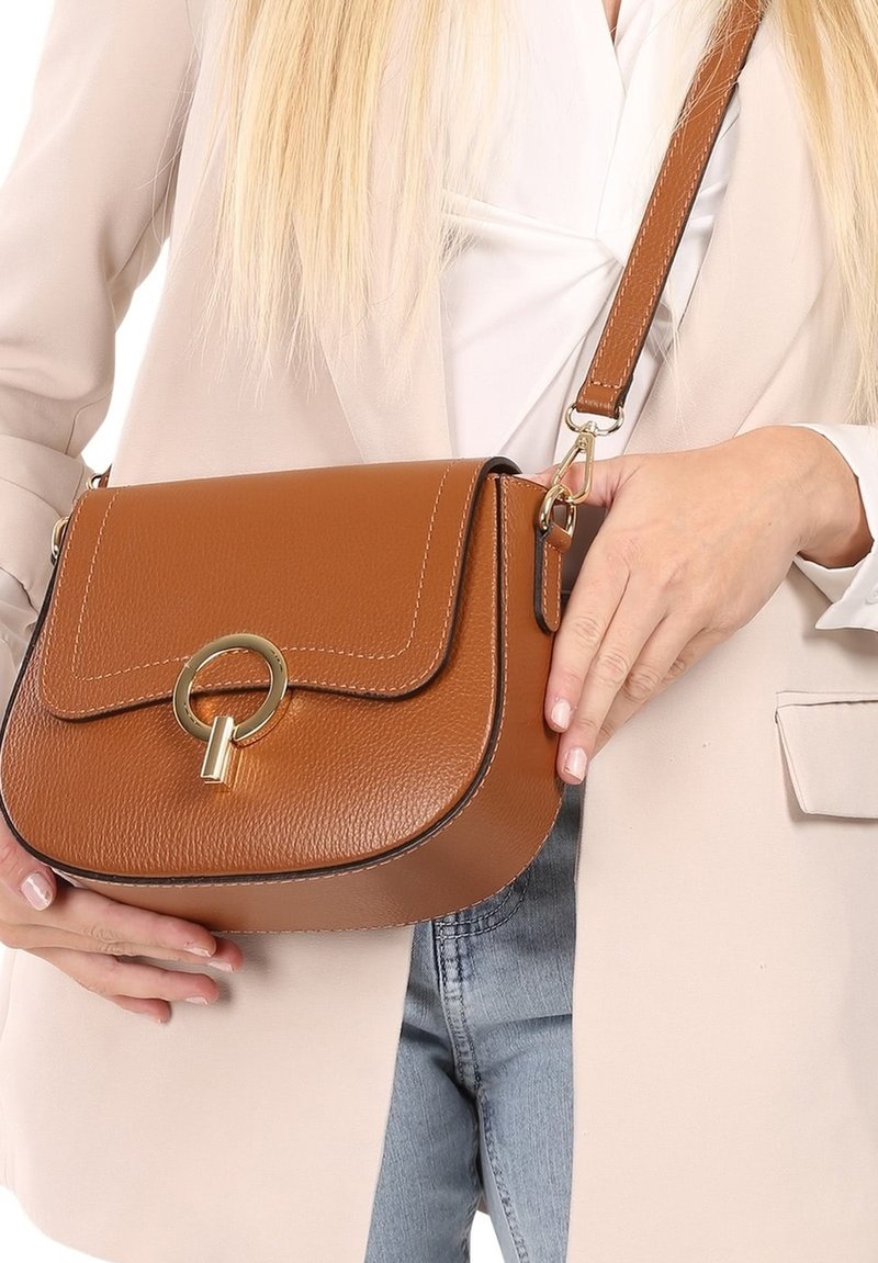 Frau trägt beige Blazer und blaue Jeans, hält eine braune Leder-Crossbody-Tasche mit goldener runder Schnalle vor einem weißen Oberteil.