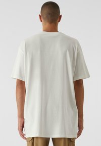 Balta per didelį t-shirt su trumpomis rankovėmis, apvaliu kaklu ir lygiu paviršiumi. Hemas gale yra ilgesnis, su šoninėmis skeltuvėmis, kad būtų patogiau.