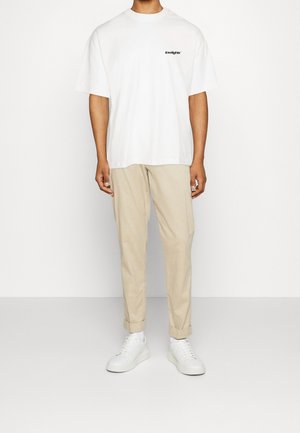 Camiseta blanca de algodón con un logo negro, combinada con pantalones chinos beige y zapatillas blancas. Diseño sencillo, corte relajado, mangas cortas, bajos de los pantalones remangados.