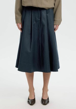 Persona che indossa una gonna midi plissettata blu navy, una camicia marrone a quadri a maniche lunghe e tacchi neri a punta, in piedi su un pavimento bianco.