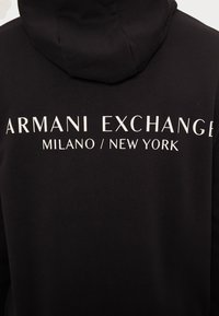 Black sweatshirt with a hood featuring the text "ARMANI EXCHANGE MILANO / NEW YORK" in large white letters on the back. Smooth texture.