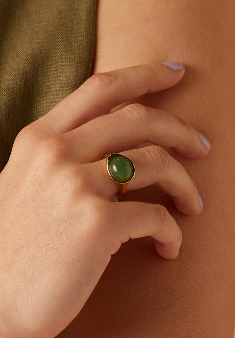 Goldring mit einem großen, ovalen grünen Stein, der in eine glatte Fassung gefasst ist. Das Band ist einfach und schmal und ruht auf einer Hand mit heller Haut.