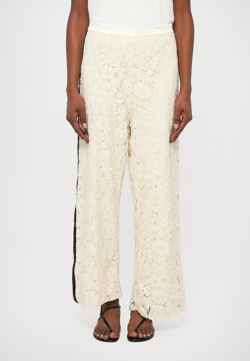 Calças largas em renda na cor creme, com um padrão floral texturizado, cintura elástica e painel lateral preto em contraste.