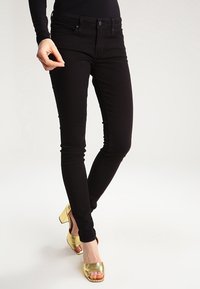 Jeans skinny negros con cinco bolsillos, detalles de costura y un ajuste ceñido. Combinados con sandalias de tacón doradas metálicas, con punta abierta y acabado texturizado.