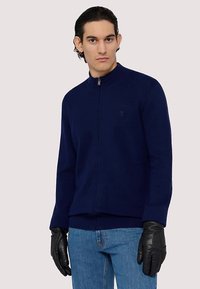 Marinblå zip-up-tröja med ribbad textur, hög krage och subtil logotypdetalj. Matchad med blå jeans och svarta läderhandskar.