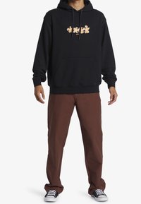 Persona con sudadera negra con capucha y diseño floral naranja, pantalones marrones y zapatillas negras y blancas de pie sobre un fondo blanco.