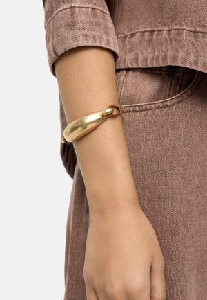 Ramię w brązowym denimowym ubraniu z gładką, masywną złotą bransoletą na nadgarstku.