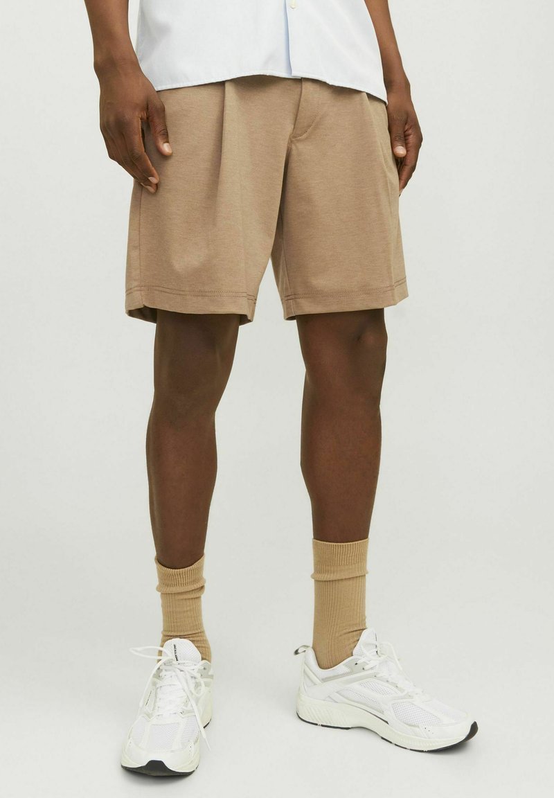Beige knit shorts with a relaxed fit, featuring a cuffed hem and side pockets. Worn with white sneakers and tan socks.