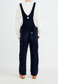 Marineblaue Corduroy-Overall mit breiten Schulterträgern, großen Fronttaschen und einem lockeren Schnitt. Getragen über einem cremefarbenen Langarmshirt.