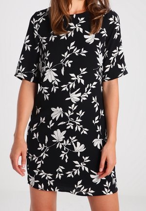 Kvinde iført en sort kortærmet kjole med et hvidt blomstret og bladmønster, stående mod en ensfarvet lys baggrund.