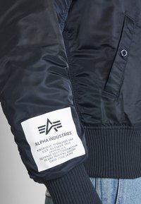 Bomba jakna tamnoplave boje s rebrastim manžetama i ovratnikom, sa bijelom etiketom koja prikazuje logo brenda i informacije o proizvodu na rukavu.