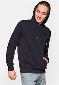 Granatowy hoodie wykonany z miękkiego materiału, z kieszenią z przodu, regulowanym kapturem na sznurki i ściągaczami, noszony z jasnoniebieskimi dżinsami.