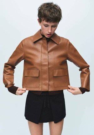 Chaqueta de cuero marrón con cuello, dos bolsillos frontales y detalles acanalados en los puños, llevada sobre una falda negra. Textura suave.