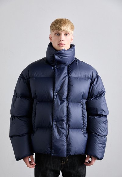 Jeune homme portant une doudoune oversized bleu marine avec un col montant, se tenant contre un fond gris clair uni.