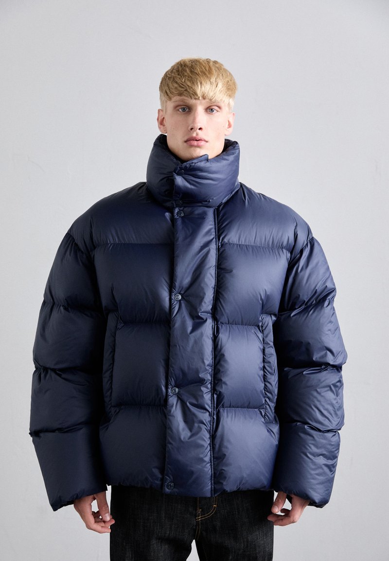 Junger Mann in einer übergroßen, marineblauen Puffjacke mit hohem Kragen, der vor einem schlichten hellgrauen Hintergrund steht.