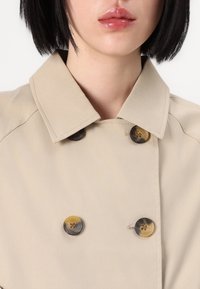 Gabardina beige con cuello estructurado, que presenta dos grandes botones en contraste y una tela suave y texturizada.