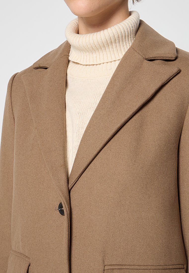 Brauner Mantel mit Reverskragen, einzelner schwarzer Knopfschluss und vorderen Taschen, getragen über einem leichten cremefarbenen Rollkragenpullover.