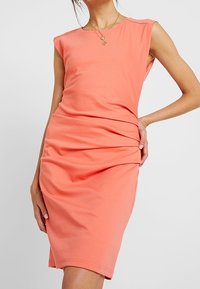 Orange ärmlös klänning i stretchigt tyg, figurnära siluett med rynkdetalj vid midjan, slät textur, knälång nederdel.