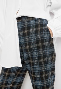 Zwarte en blauwe geruite broek van een gestructureerde stof, met dunne bruine lijnen, gecombineerd met een losse witte blouse.