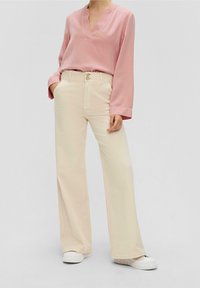 Blusa rosa chiaro a maniche lunghe con scollo a V, abbinata a pantaloni larghi crema e sneakers bianche. Tessuto morbido, vestibilità rilassata.