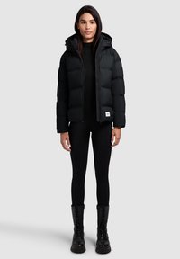 Sort pufferjakke med hætte, med frontlynlås, elastiske manchetter og en hvid logo-patch. Sammen med sorte leggings og kampstøvler.
