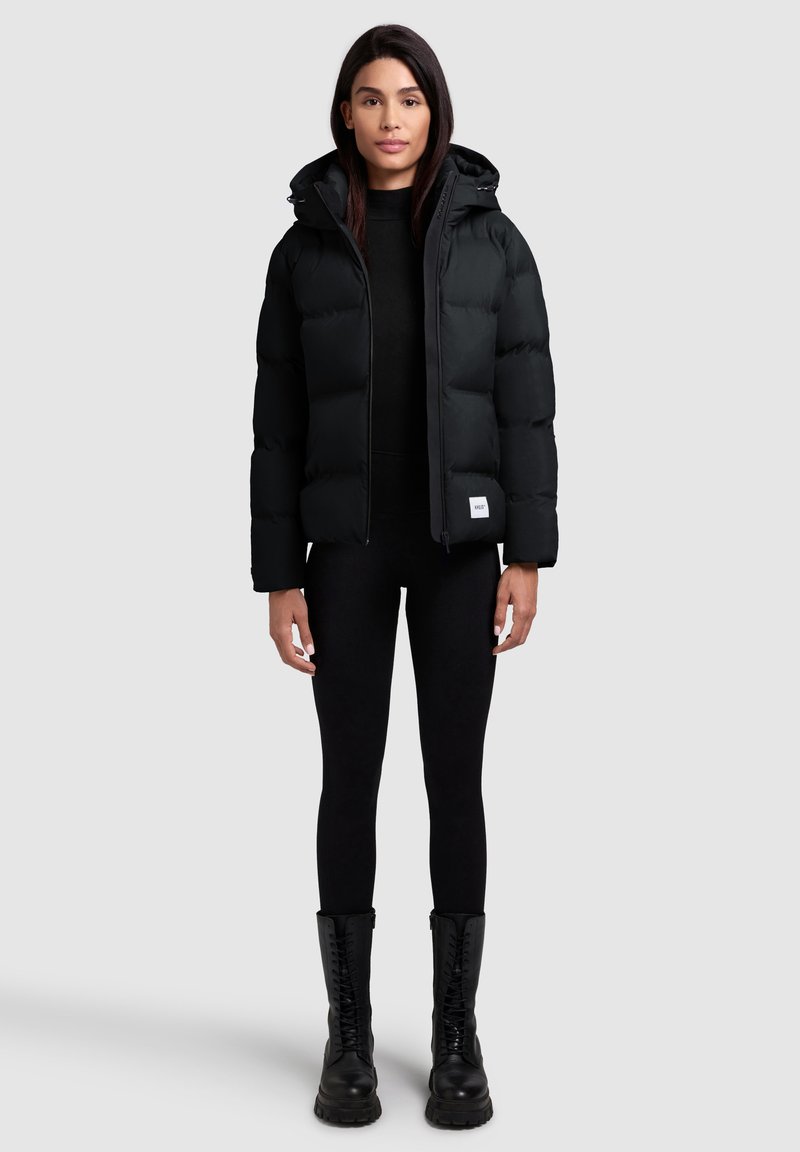 Sort pufferjakke med hætte, med frontlynlås, elastiske manchetter og en hvid logo-patch. Sammen med sorte leggings og kampstøvler.