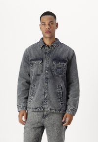 Chaqueta de mezclilla en gris oscuro desgastado con cierre de botones, dos bolsillos en el pecho y detalles de costura. Tejido texturizado con un aspecto ligeramente usado.