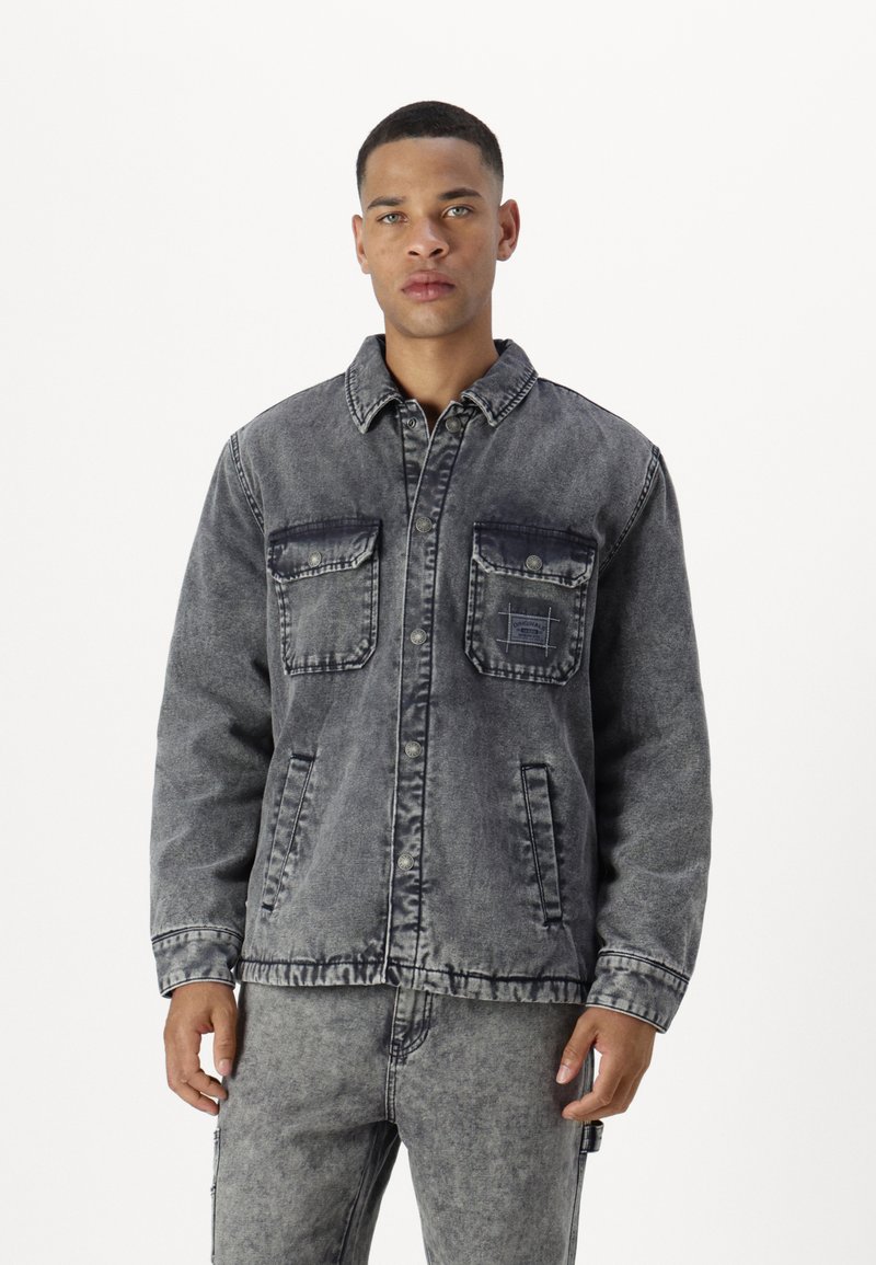 Chaqueta de mezclilla en gris oscuro desgastado con cierre de botones, dos bolsillos en el pecho y detalles de costura. Tejido texturizado con un aspecto ligeramente usado.