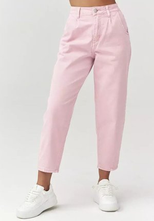 Kobieta ubrana w jasnoróżowe, krótkie spodnie z wysokim stanem oraz białe trampki, stojąca z lekko rozstawionymi stopami na jednolitym tle.