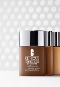 Maquillaje líquido Clinique Anti-Blemish Solutions en un frasco de vidrio marrón con tapa plateada, presenta un etiquetado limpio sobre un fondo sutil con un patrón de puntos.