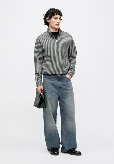 Sivá vzorovaná bunda na zips s vysokým golierom, v kombinácii so širokými vyblednutými modrými džínsami s rozšírenými nohavicami. Čierna kabelka a kožené topánky dotvárajú vzhľad.