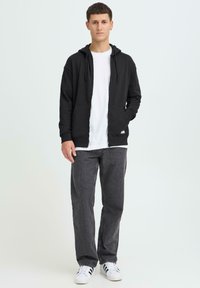 Sweat à capuche noir zippé avec poches avant, porté sur un t-shirt blanc. Associé à un jean gris foncé et des baskets blanches avec des accents noirs.