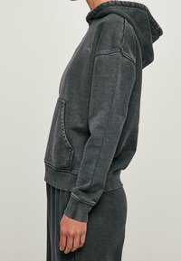 Vista laterale di una persona che indossa un hoodie nero sbiadito e pantaloni della tuta abbinati, con la tasca frontale dell'hoodie visibile e un fit rilassato.