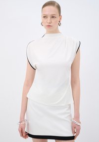 Top bianco con maniche corte strutturate e rifiniture nere, abbinato a una gonna bianca coordinata con orlo nero. Bracciale circolare trasparente come accessorio.