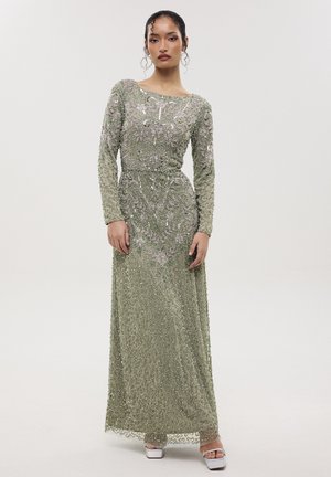 Kobieta o ciemnych włosach, ubrana w długą, sięgającą podłogi, srebrno-zieloną suknię wysadzaną koralikami z długimi rękawami oraz białe sandały na platformie, stojąca na jednolitym tle.