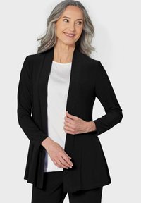 Schwarze Strickjacke mit offener Vorderseite und langen Ärmeln, aus weichem Material gefertigt. Kombiniert mit einem weißen Oberteil, das in der Nähe des Ausschnitts dezente Glitzereffekte aufweist.