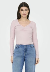 Langærmet, lys pink ribbet top med V-hals og rynket detalje foran. Kombineret med højtaljede blå denimjeans.