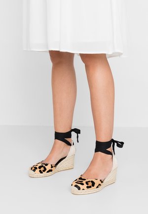 Jambes portant des espadrilles compensées à imprimé léopard sur l'avant, rubans noirs s'enroulant autour des chevilles, et jupe blanche se terminant au-dessus des genoux sur un fond neutre.