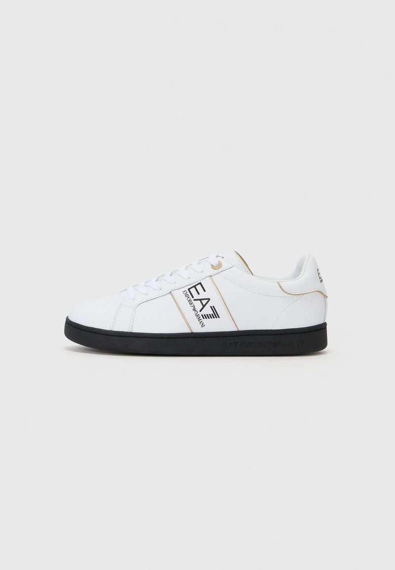 Λευκά αθλητικά παπούτσια με μαύρη καουτσούκ σόλα. Διαθέτουν χρυσές λεπτομέρειες και σήμα στο πλάι. Το υλικό φαίνεται λείο και συνθετικό.