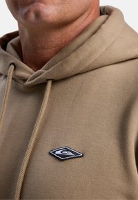 Beige hoodie met een textuurafwerking, een trekkoordcapuchon en een zwart-wit geborduurd logo op de linkerborst.