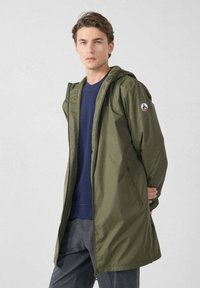 Giacca con cappuccio verde oliva con chiusura a zip frontale, due tasche laterali e patch con logo sulla manica. Leggera, con texture liscia, design di media lunghezza.